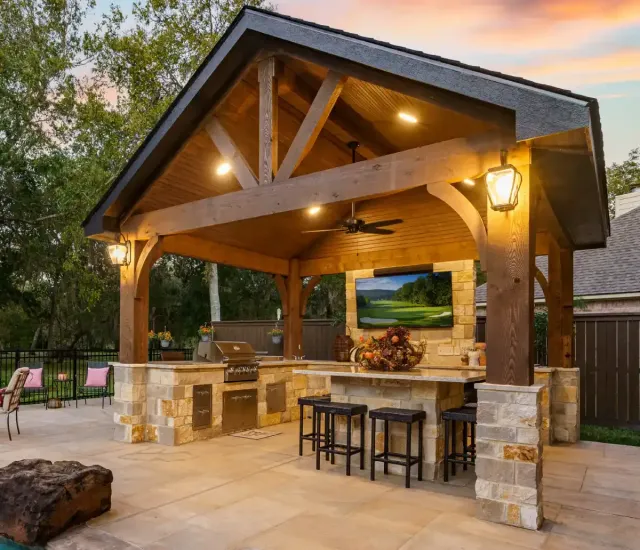 Covered outdoor kitchen with stone bar, wooden beams, and warm lights at dusk