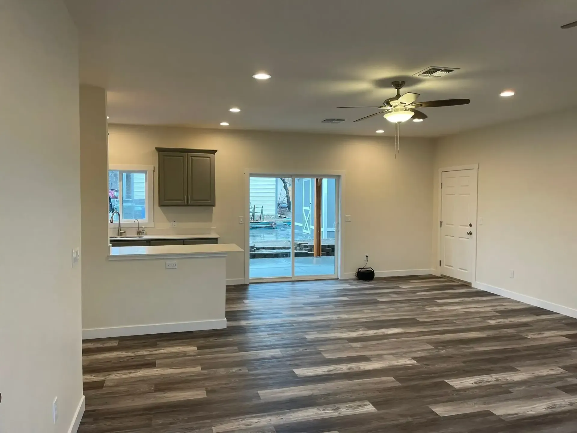 Empty open-plan living room with kitchen, wood-look flooring, ceiling fan, and sliding glass door.