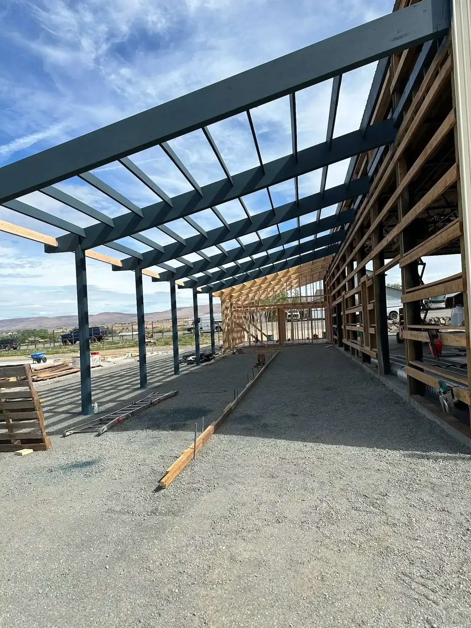 Steel frame building under construction on a gravel site, with open beams and partial wooden scaffolding.