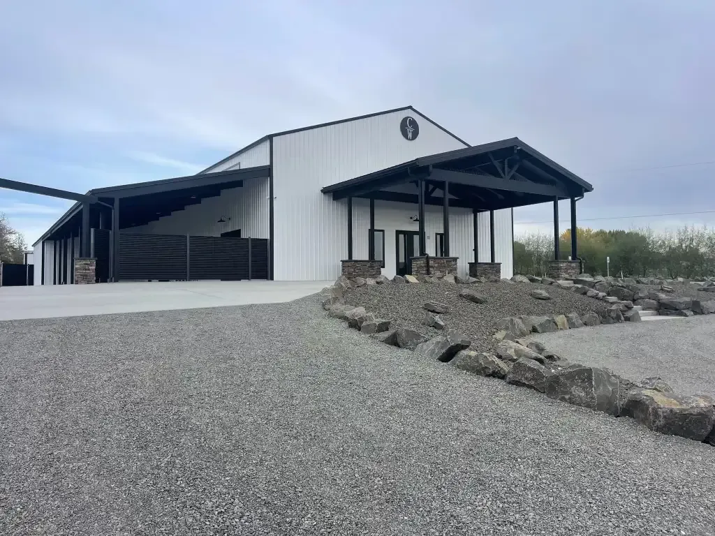 White metal barn-style building with black roofed porches, gravel driveway, and rocky landscaping.