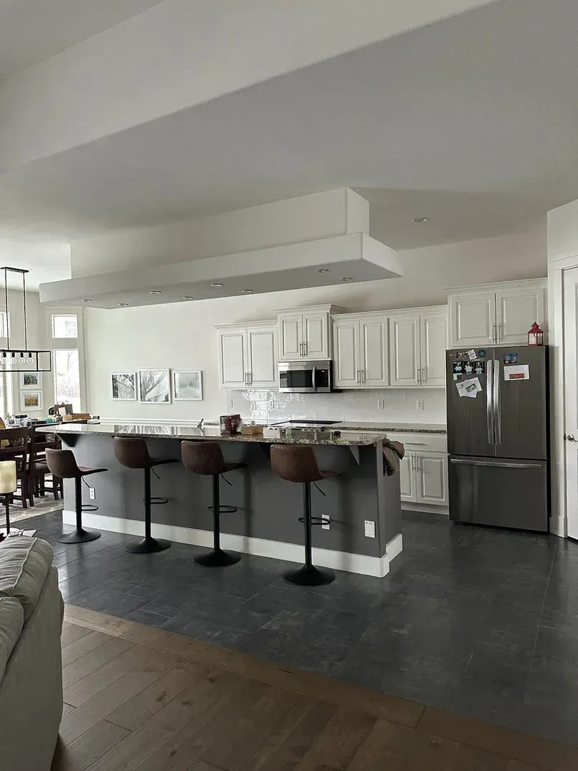 Modern kitchen and open living area with white cabinets, gray floors, island stools, and a stainless steel fridge