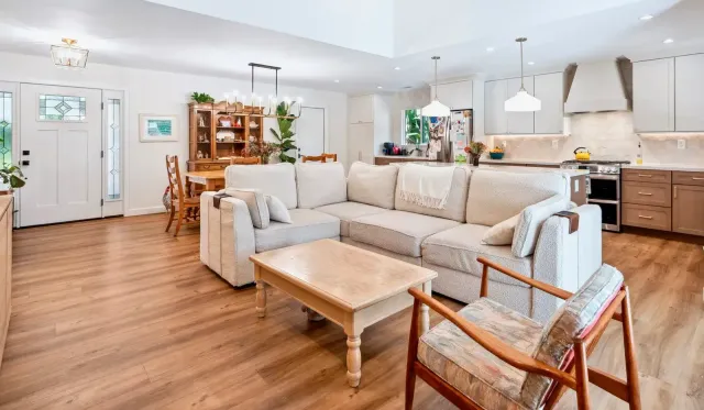 Bright open-concept living room with beige sectional, wood floors, dining area, and kitchen in the background