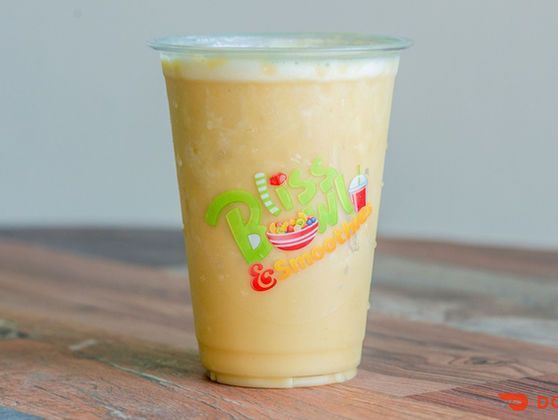 Plastic cup of a pale yellow drink with foam on top, sitting on a wooden table.