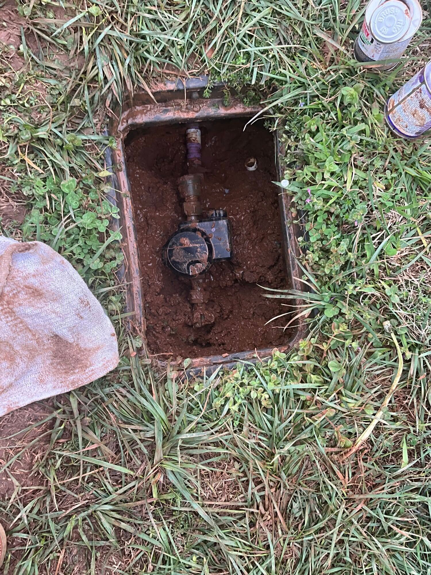 Open utility box with a valve inside, set in muddy ground and grass.