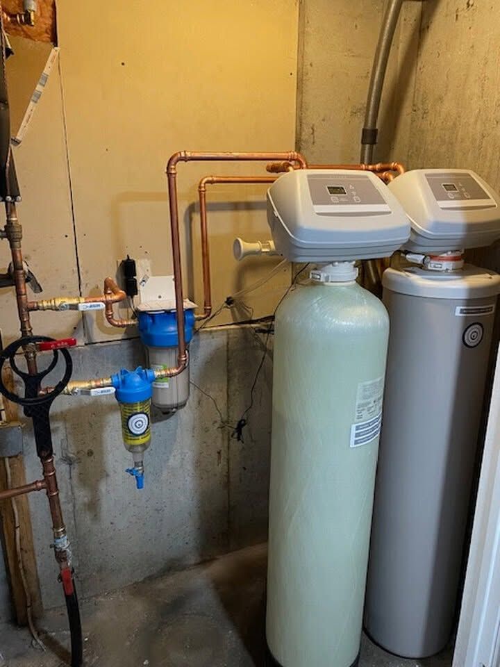 Water softener and filtration tanks in a basement utility area with exposed pipes.