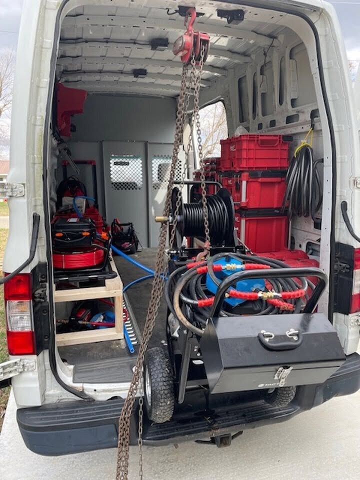 Inside a white van, a red cable reel and coiled hoses on a black equipment cart with tools and storage bins.