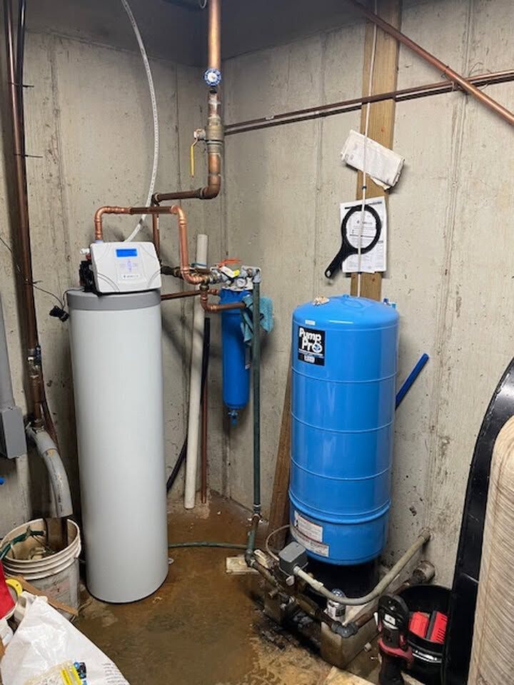Basement utility area with white and blue water treatment tanks, pipes, and gauges.