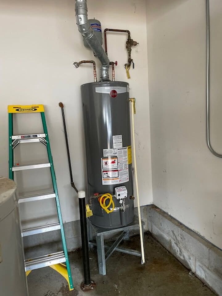 Water heater in a utility room beside a ladder, with exposed pipes and a concrete floor.