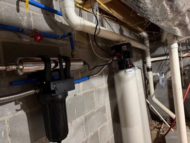 Plumbing and filtration equipment in a basement, with pipes, valves, and a white water filter housing.
