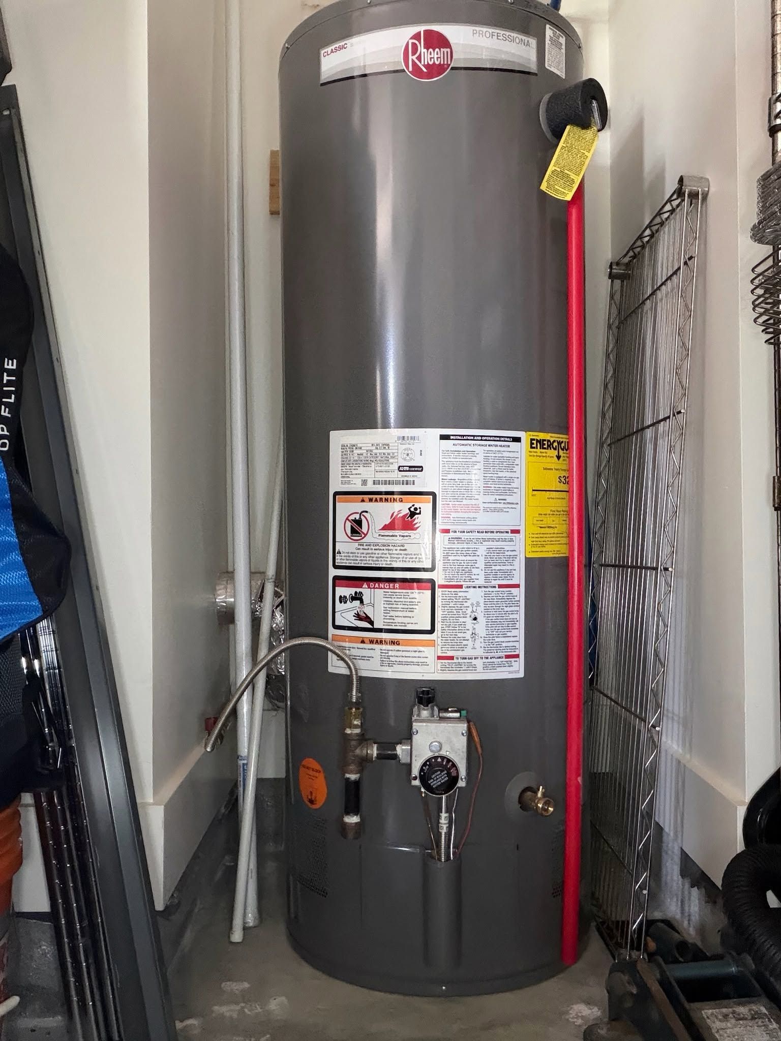 Tall gray water heater in a narrow utility closet with labels, pipes, and a red hose attached