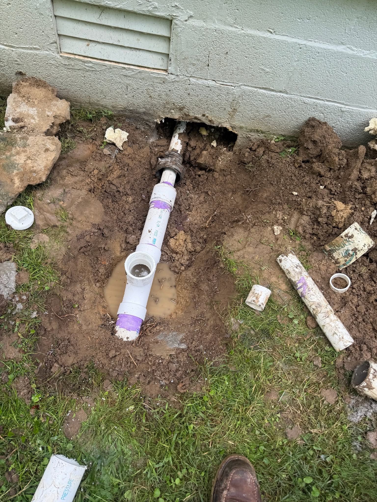 Exposed white PVC pipe in a dirt trench beside a concrete wall, with tools and pipe pieces nearby.