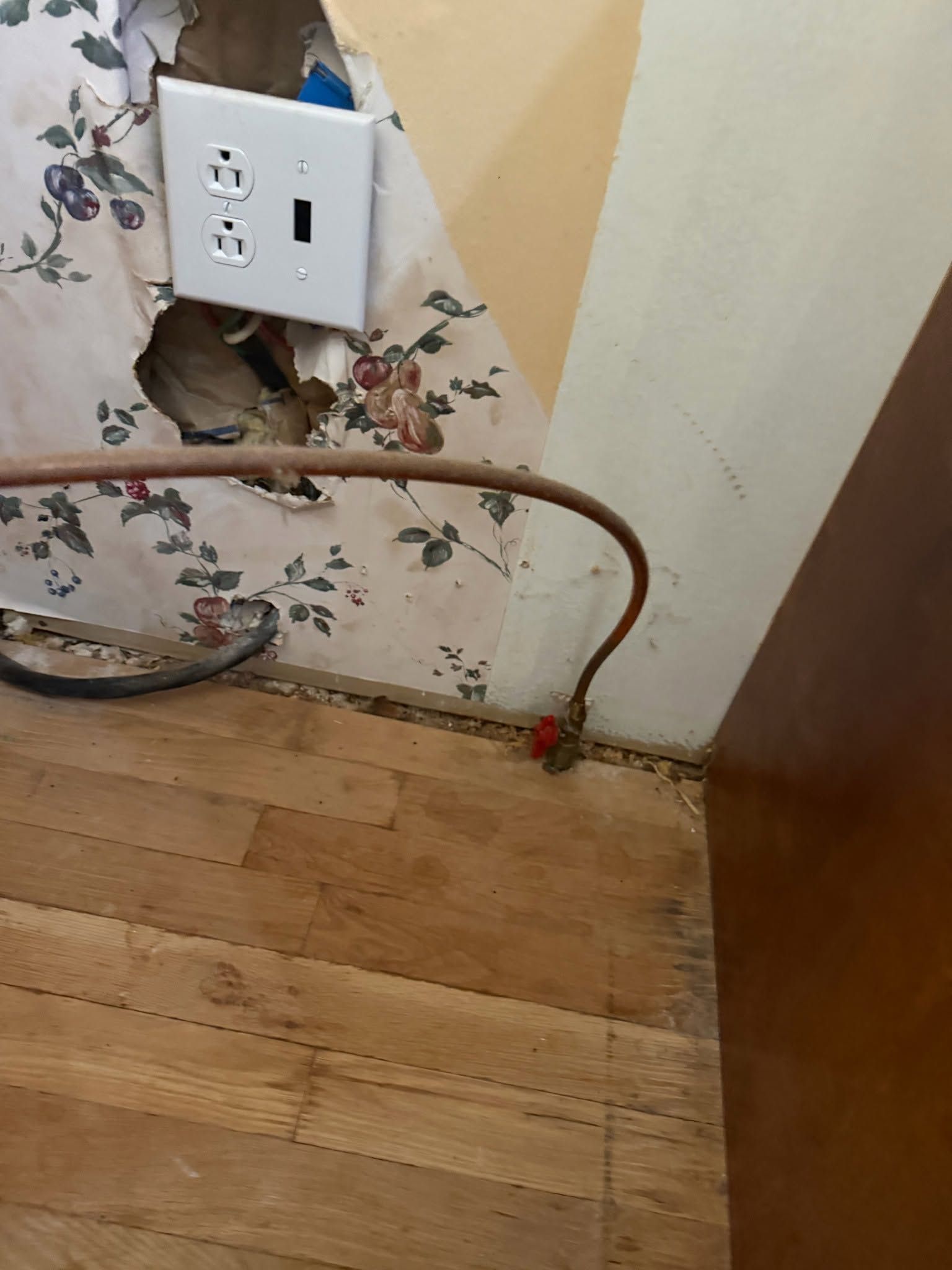 Damaged wall outlet with exposed wiring near a hardwood floor and baseboard corner