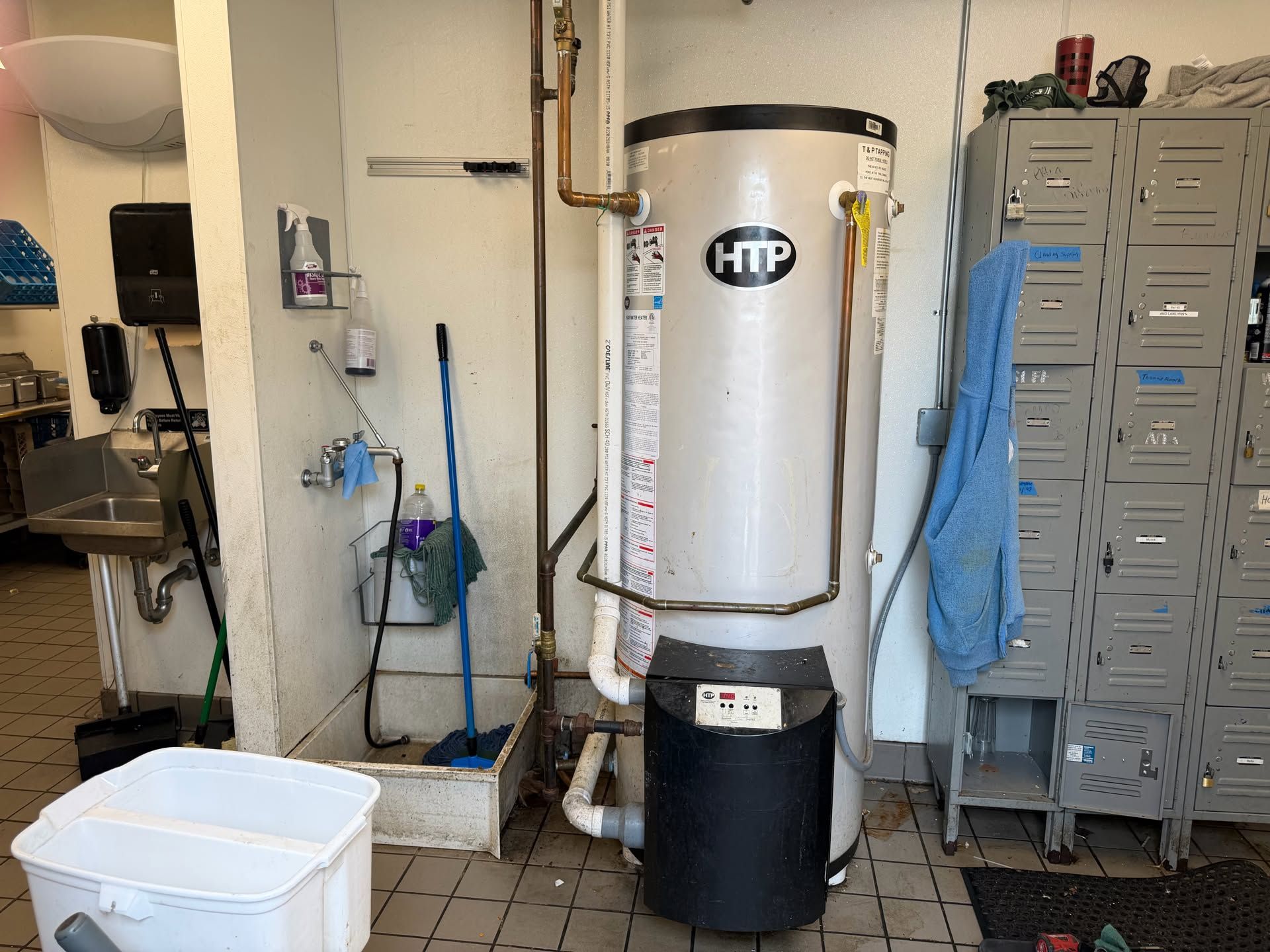 Water heater in a utility room beside gray lockers, with a white bin and cleaning items nearby.