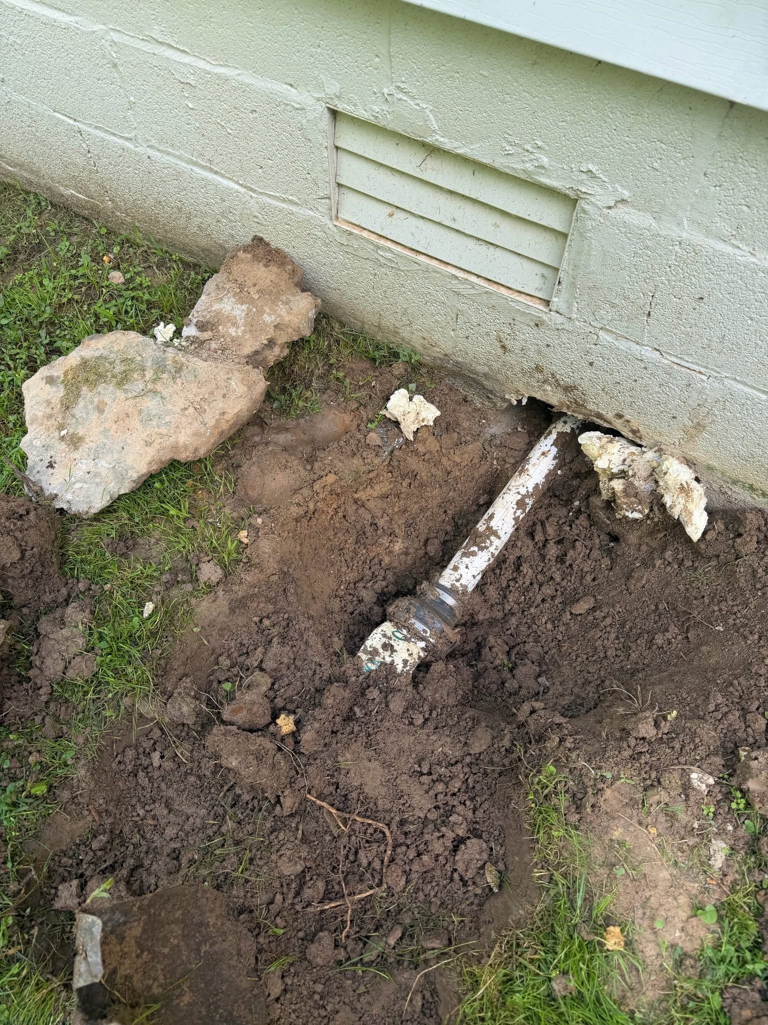 Damaged lawn beside a building wall with rocks and exposed pipe or root in muddy soil