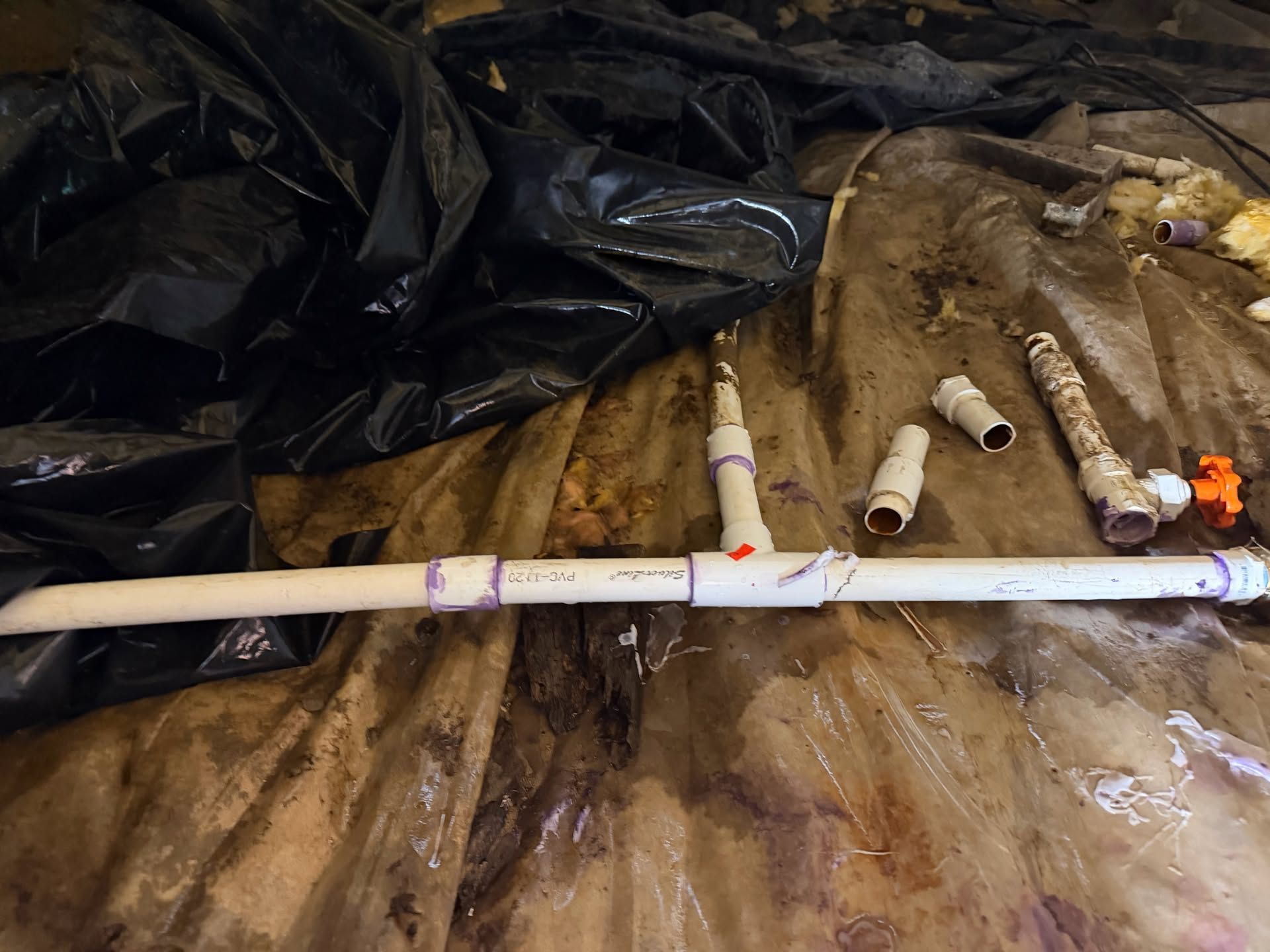 White PVC pipe sections and fittings scattered on brown tarp beside a black plastic sheet.