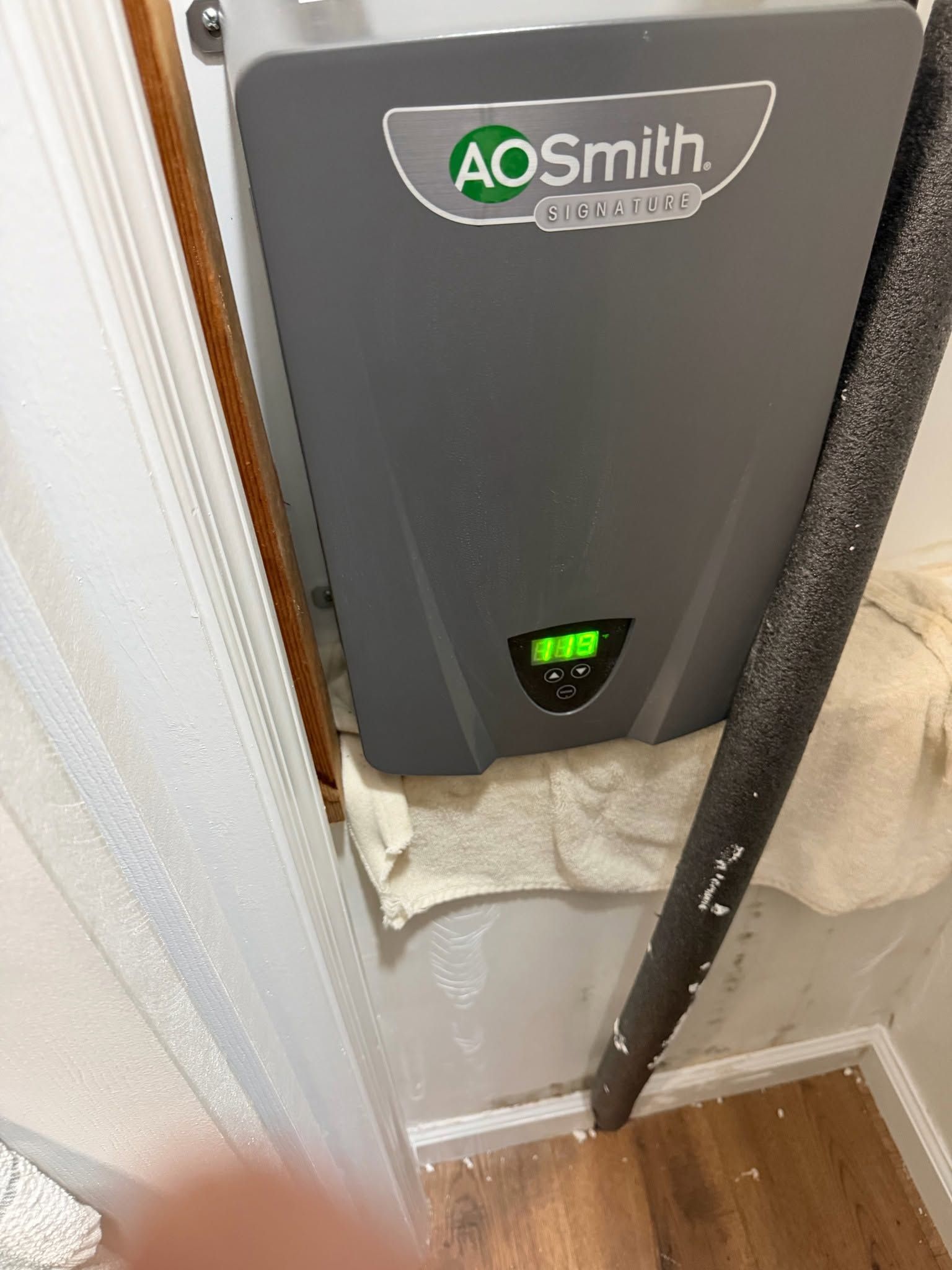 AO Smith wall-mounted water heater in a narrow closet, gray unit with green display and pipe below