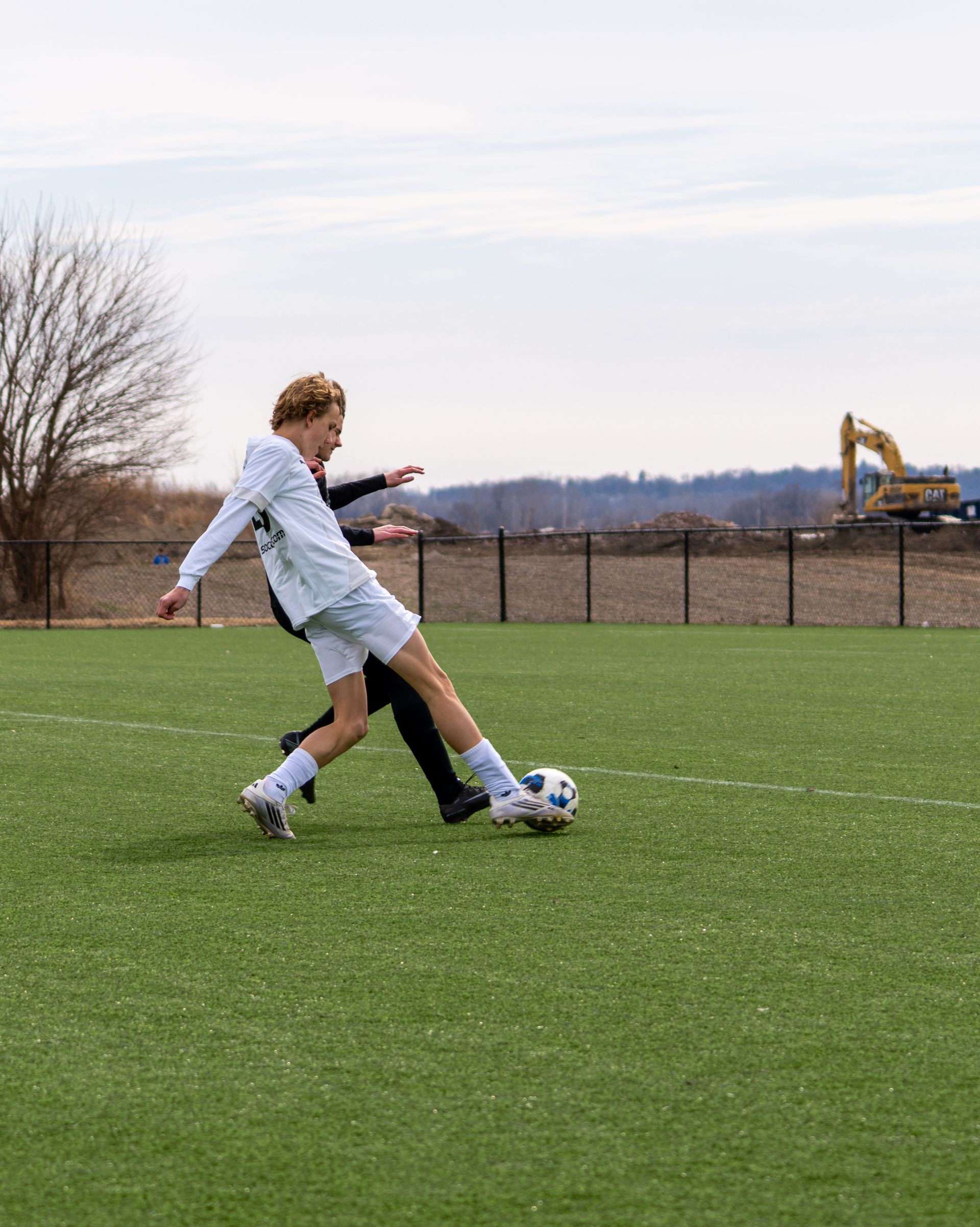 Soccer player in white jersey kicking a ball on a grassy field with an excavator in the background