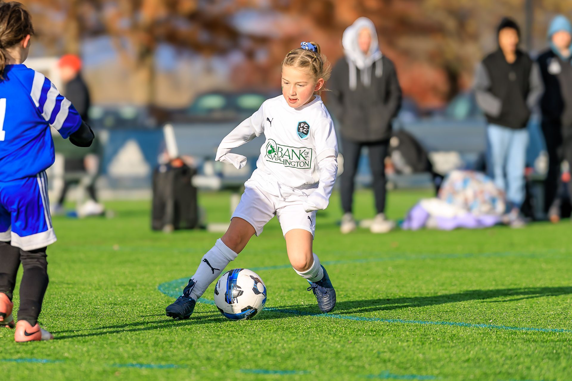 Child in white soccer uniform dribbles a ball on a grassy field during a youth game