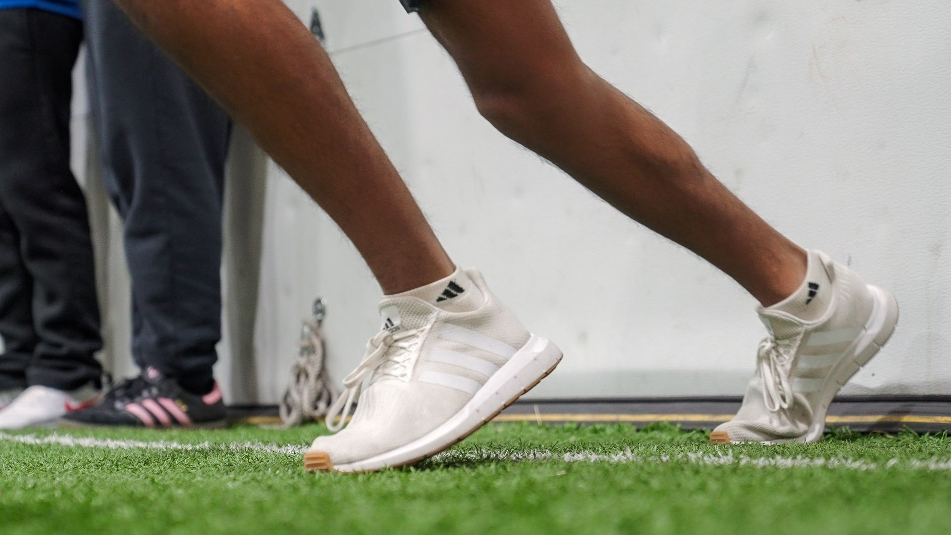 Runners’ legs in white sneakers on a green field, one mid-stride during practice