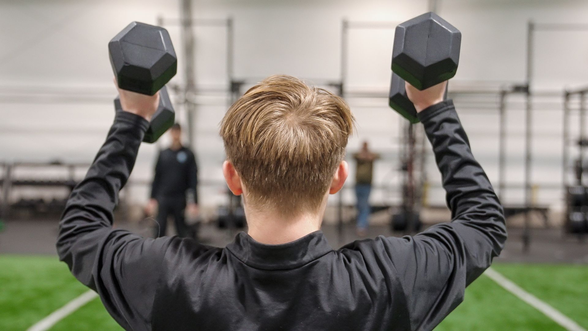 Person lifting dumbbells overhead in a gym, seen from behind on a turf floor