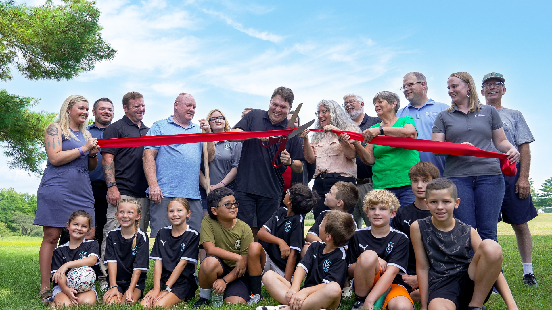 Group of people outdoors at a ribbon-cutting ceremony, holding a red ribbon and smiling on a grassy field.