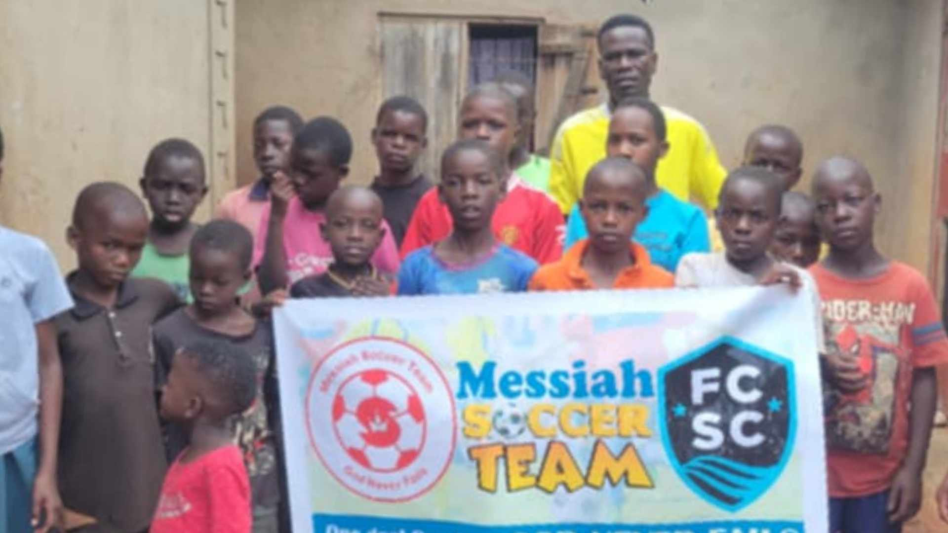 Children and two adults pose with a banner reading “Messiah Gecen Team” and “FC SC”