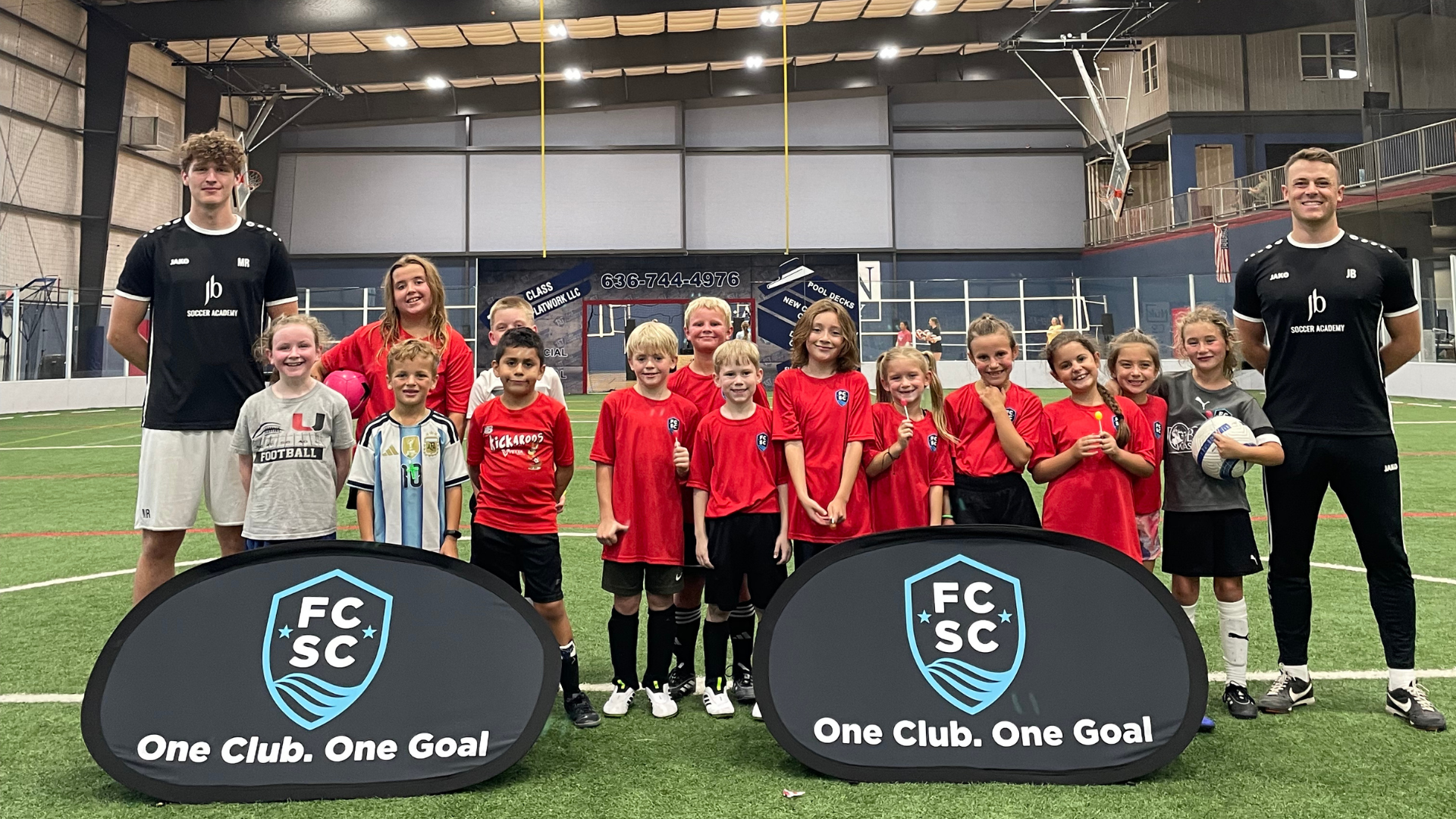 Youth soccer team posing indoors with FC SC signs, two coaches, and a soccer ball in the center