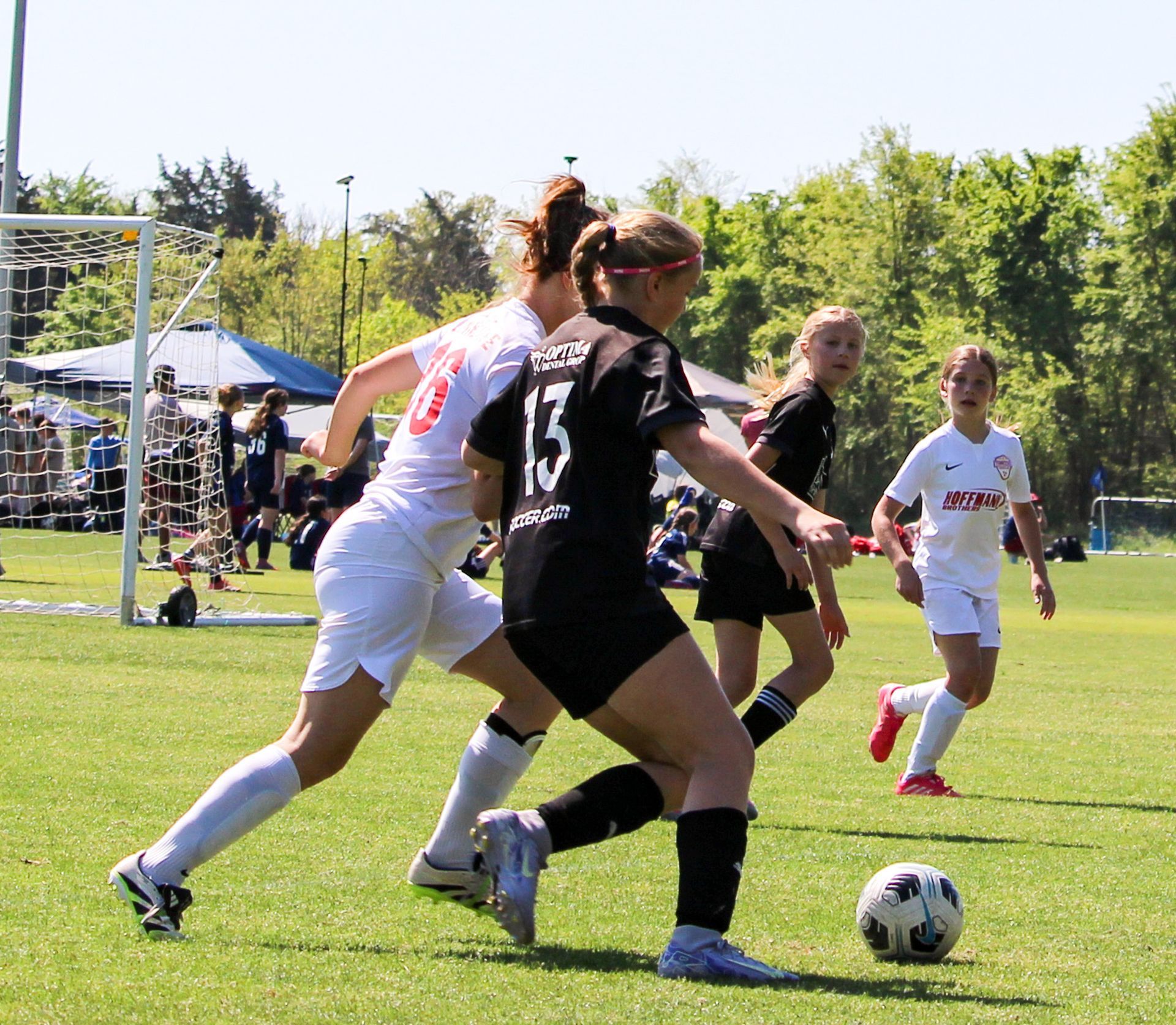 Players contest the ball during a soccer match on a grassy field, with goal and trees in the background.
