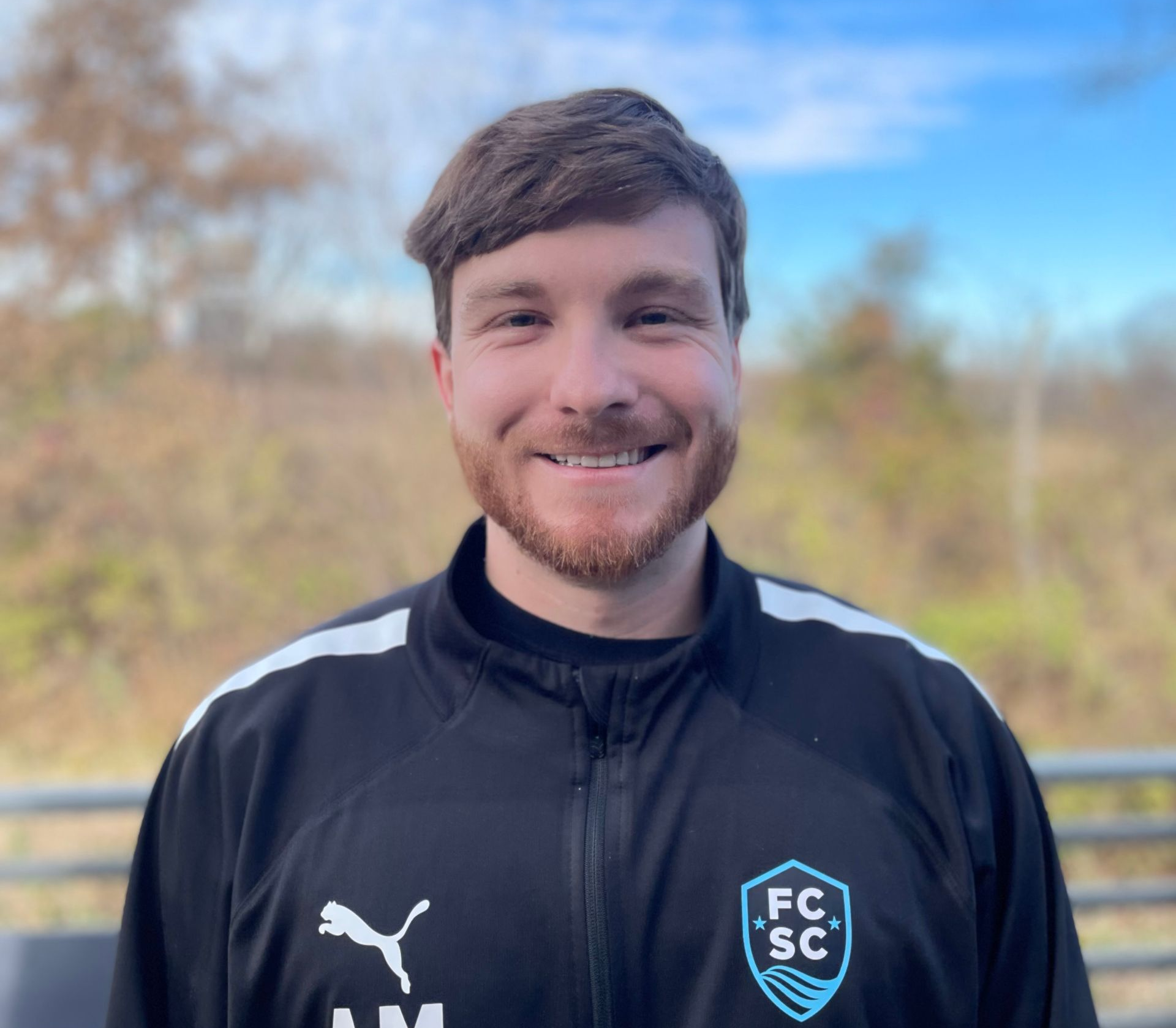 Smiling person in a black Puma FCSC jacket standing outdoors with trees in the background