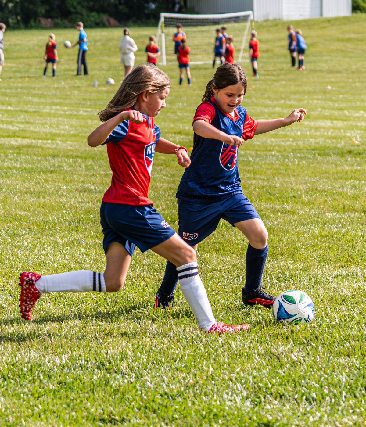 Two children in red and blue soccer uniforms chasing a ball on a grassy field