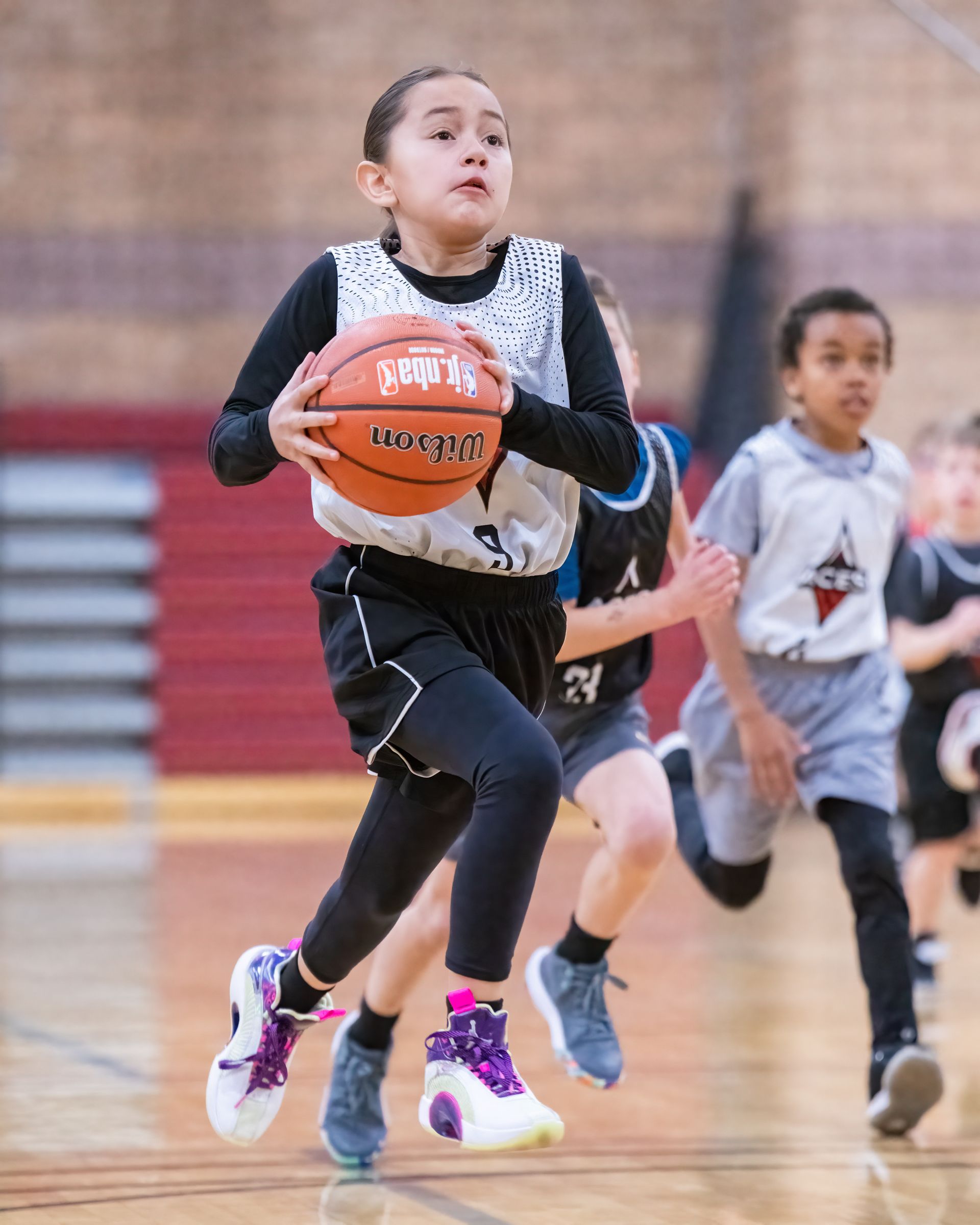 Two children sprint in a basketball game, one dribbling an orange ball on a gym court.