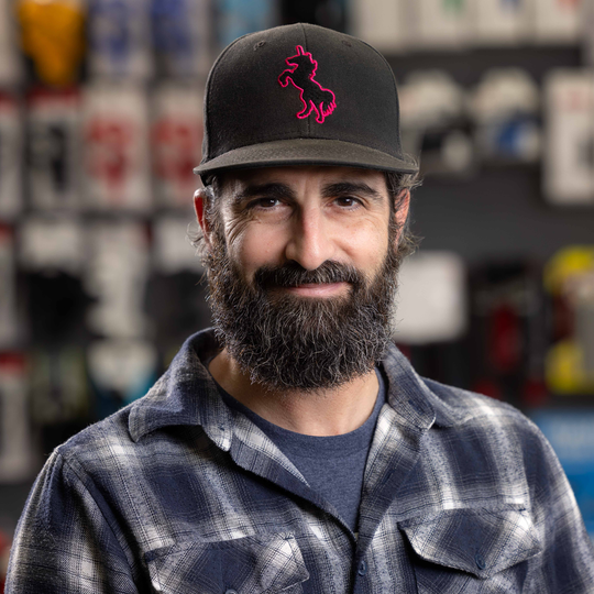 Kevin Horschel (Service Manager)-Man with beard and black cap with pink unicorn logo, wearing a plaid shirt, smiling indoors.