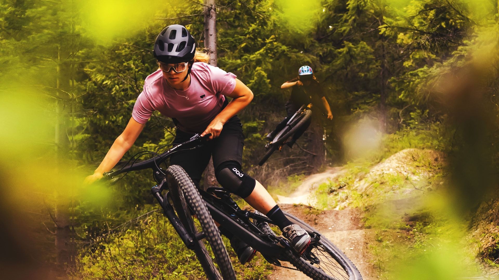 Woman on mountain bike riding trail, another biker in background.