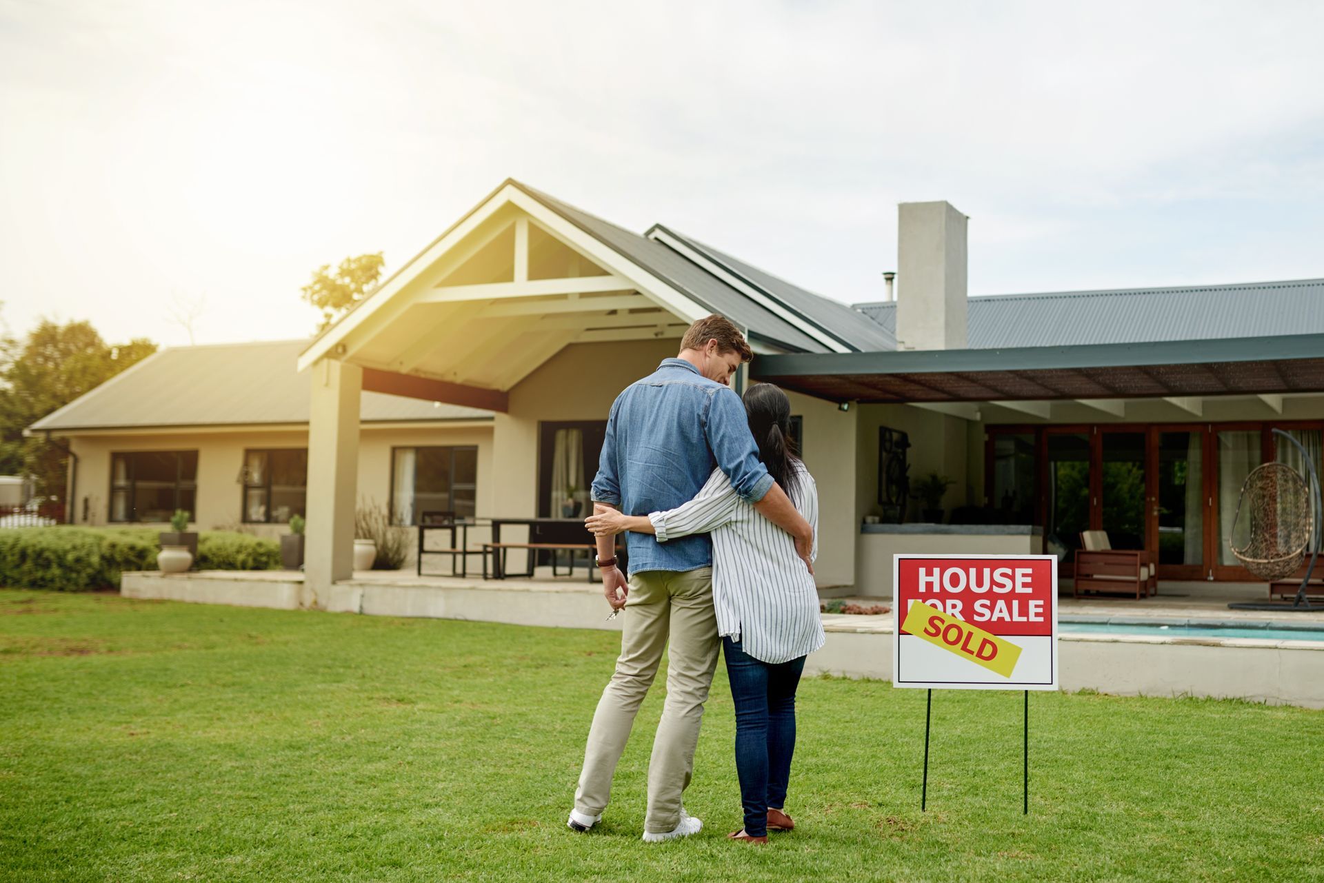 Couple stands outside a house for sale, looking at the home and sign on a lawn.