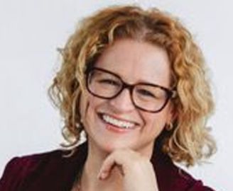 Woman with curly blonde hair and glasses smiles, resting chin on hand.