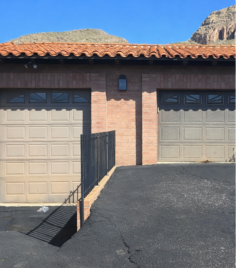 Two beige garage doors on a brick house with a black asphalt driveway and tiled roof under blue sky