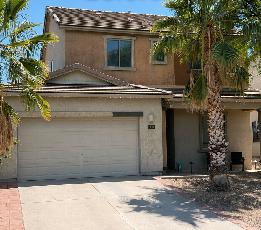 Two-story tan house with a garage, palm trees, and a sunlit driveway