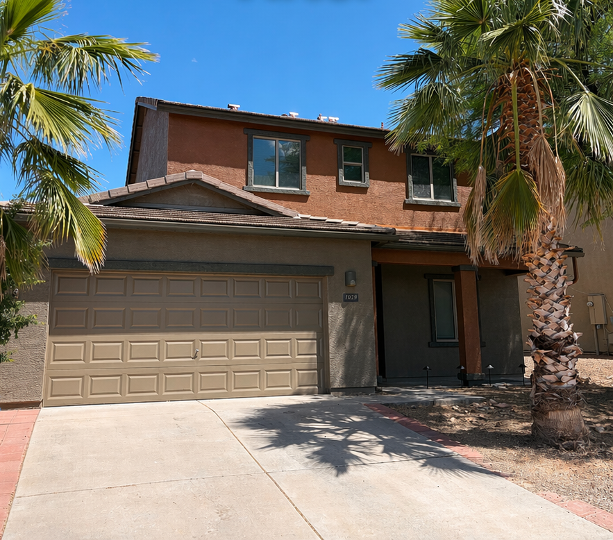 Two-story suburban house with a tan garage, concrete driveway, and palm trees in front.