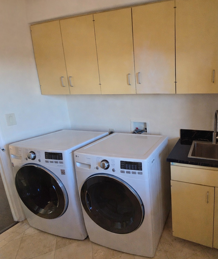 Laundry room with two front-loading washers beneath beige cabinets and a sink counter.