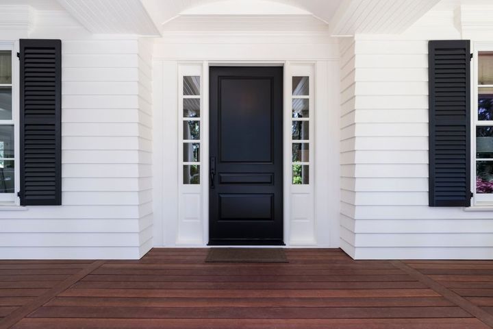 Black front door centered on a white house porch with black shutters and wood decking