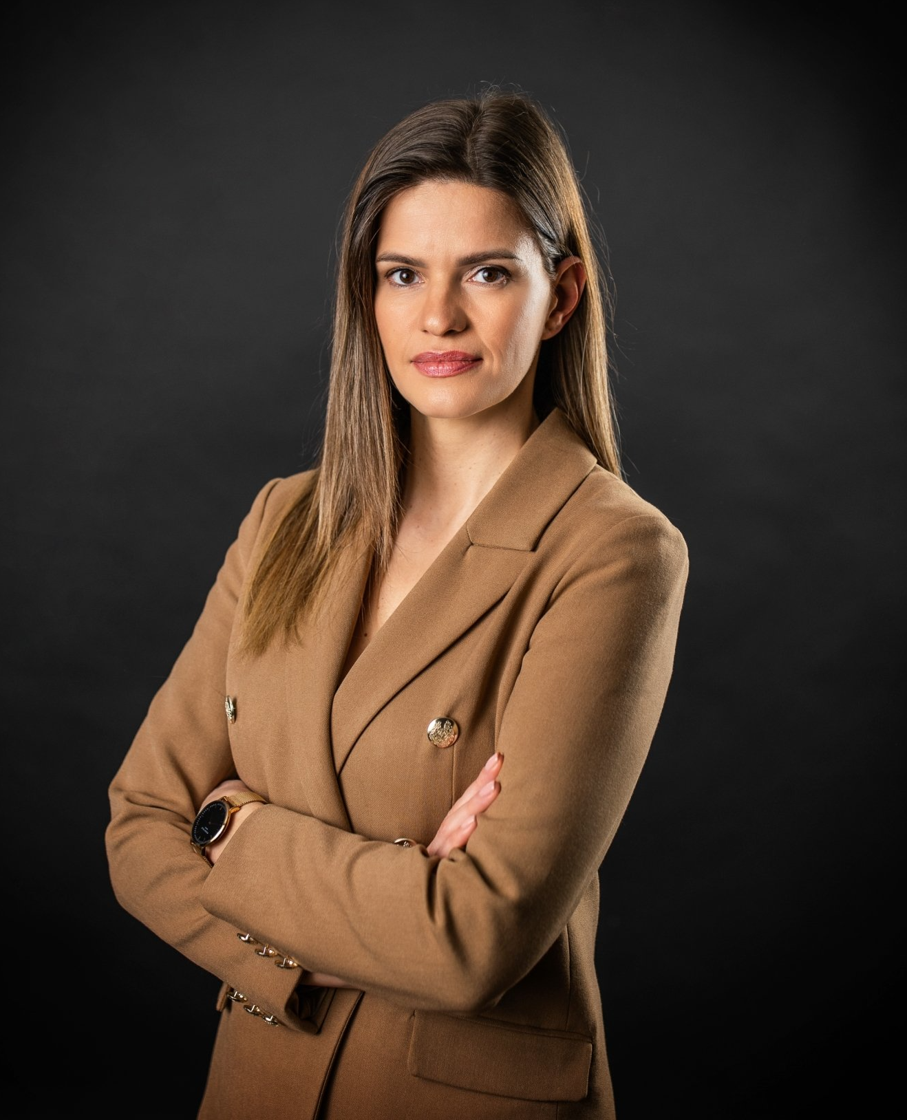 Professionelle Frau in einem beigen Blazer, die mit verschränkten Armen vor einem dunklen Hintergrund steht.
