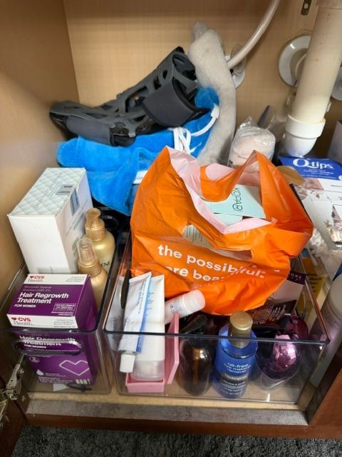 Cluttered bathroom shelf with toiletries, tissue boxes, bags, and cleaning items