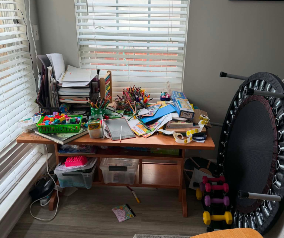 Cluttered craft desk by window, covered with papers, markers, and toys, with a mini trampoline beside it
