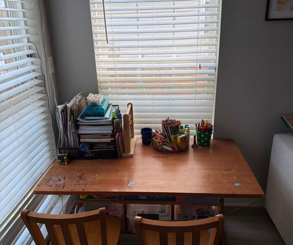 Wooden desk by windows with office supplies, pens, and papers in a bright corner workspace.