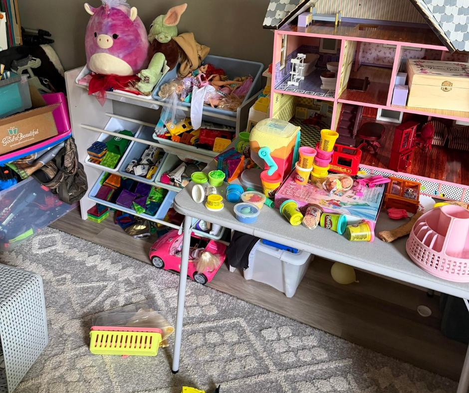 Cluttered playroom with toy tables, dollhouse, colorful toys, and stuffed animals on a rug