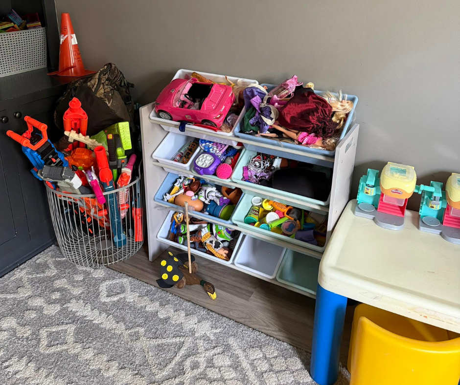 Children’s toys and bins in a corner play area, with shelves of dolls and toy vehicles on a rug.