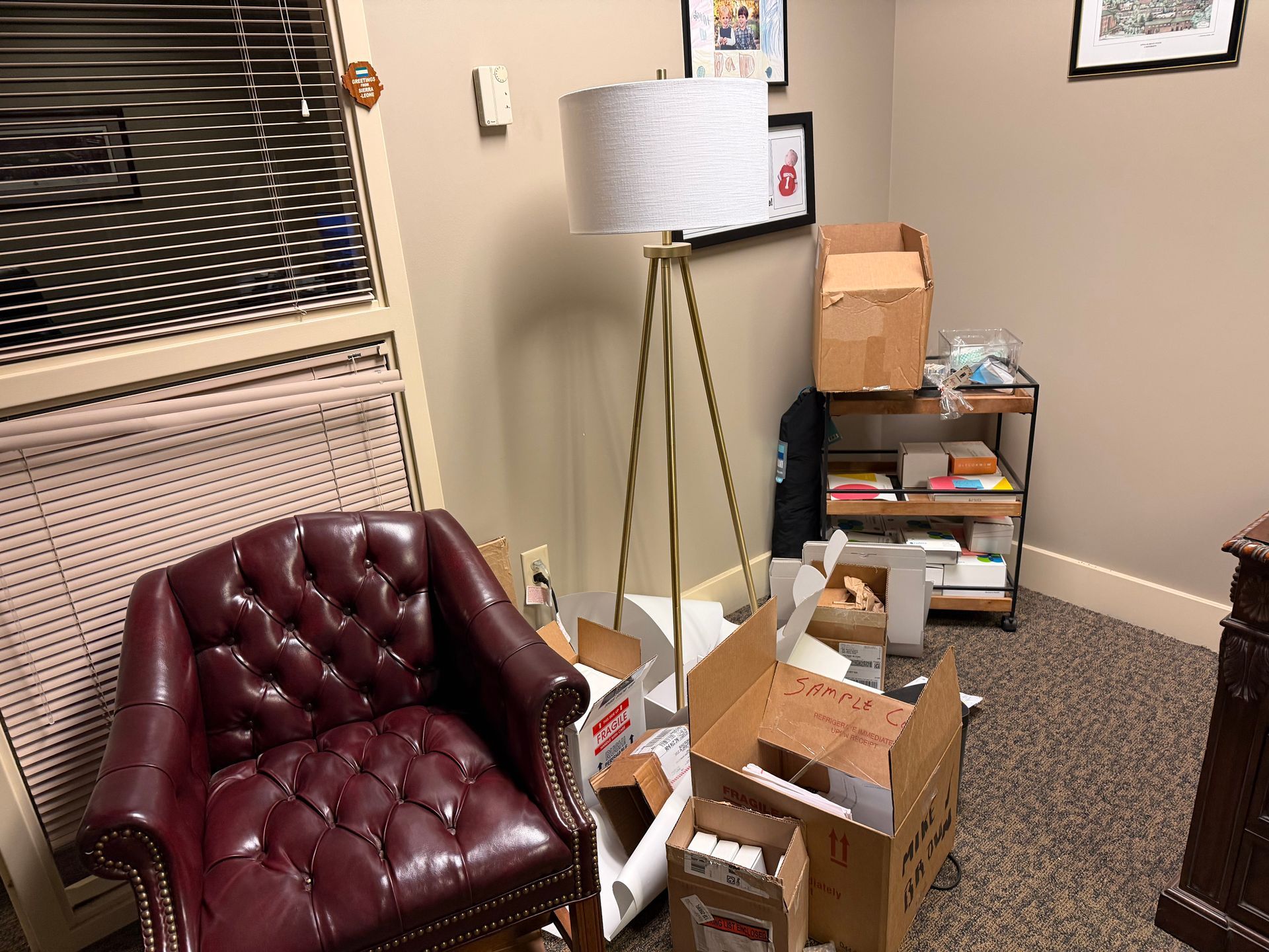 Small office corner with a burgundy chair, floor lamp, window, and stacked moving boxes.