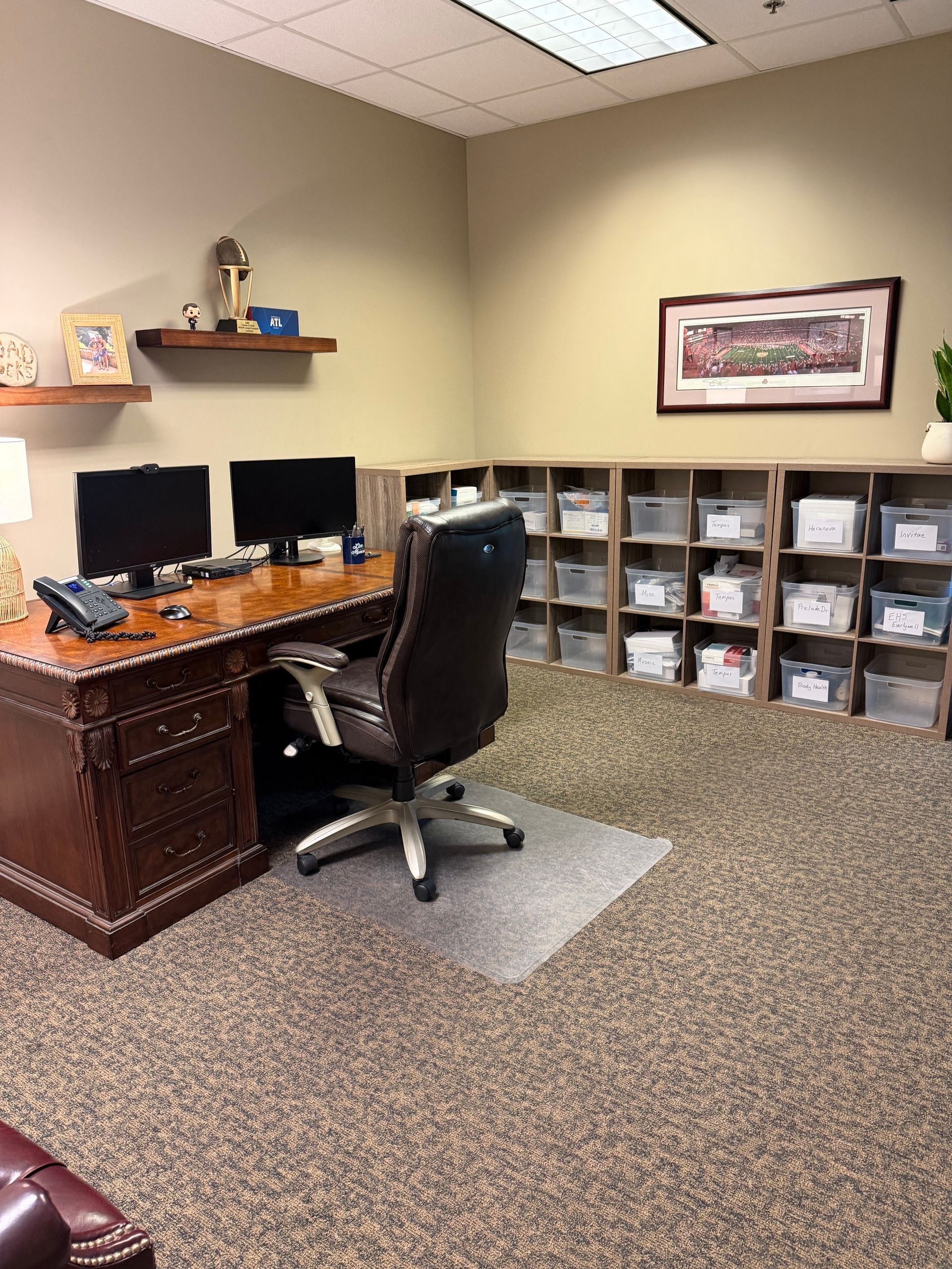 Small office with a wooden desk, two monitors, office chair, shelves of labeled bins, and framed wall art.