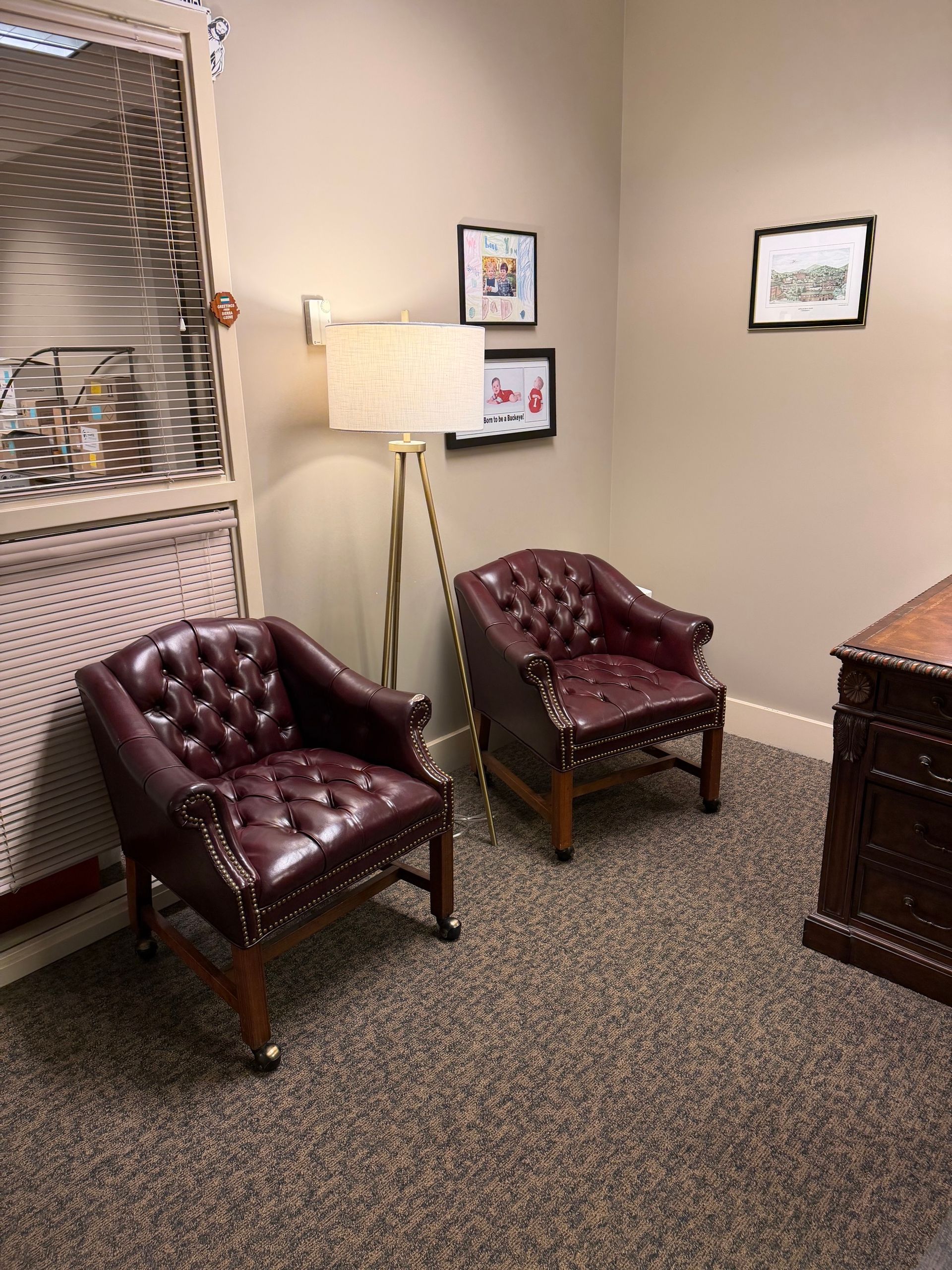 Small waiting area with two burgundy leather chairs, a floor lamp, framed art, and a window with blinds.