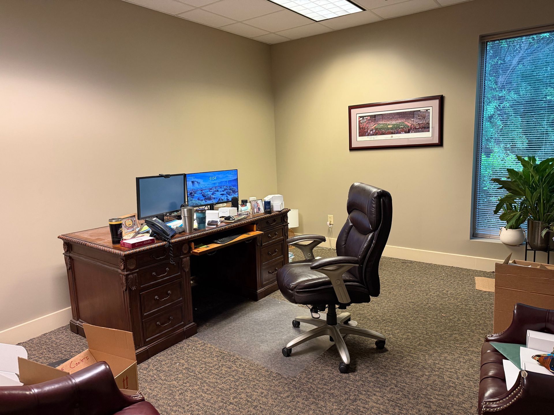 Empty office with desk, dual monitors, black swivel chair, beige walls, and a window with blue curtains.